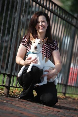 Photo of Sheila D'Arpino, smiling, kneeling by a fence, holding a small brown and white dog